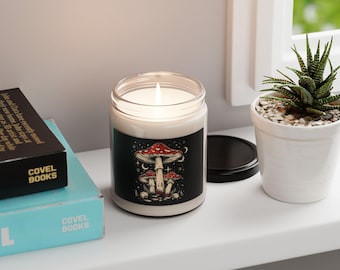 May include: A lit candle in a clear glass jar with a black label featuring a mushroom design. The mushrooms have red caps and are set against a starry background. The candle sits on a white surface next to a small potted plant.