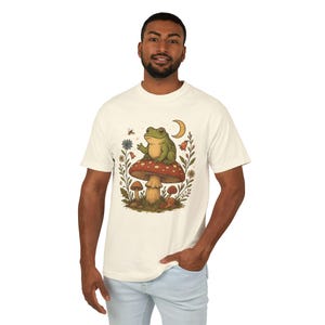 May include: Cream-colored t-shirt featuring a whimsical illustration of a green frog sitting atop a red and white mushroom. The design includes a crescent moon, flowers, and other forest elements. The shirt is made of a soft, comfortable material.