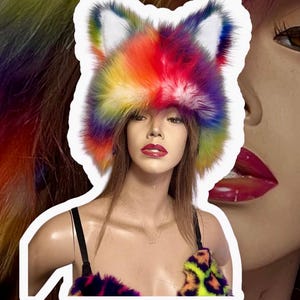 Peut inclure: Un chapeau en fausse fourrure en forme d'oreilles de renard aux couleurs de l'arc-en-ciel, rouge, orange, jaune, vert, bleu et violet. Le chapeau est porté par un mannequin aux longs cheveux bruns et au rouge à lèvres rouge. Le mannequin porte également un haut assorti.