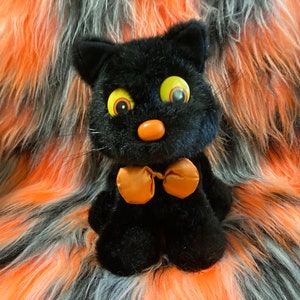 May include: Black plush cat toy with large yellow eyes and an orange bow tie. The cat is sitting on a black, orange, and white fuzzy surface.
