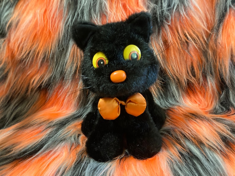May include: Black plush cat toy with large yellow eyes and an orange bow tie. The cat is sitting on a black, orange, and white fluffy surface.