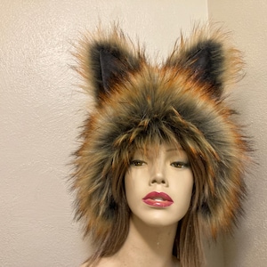 May include: A faux fur fox hat with black, brown, and orange coloring. The hat features upright ears and a fluffy texture, designed to resemble a fox. The hat is worn by a mannequin.
