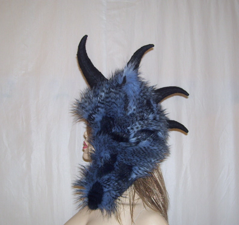 May include: A blue and black faux fur hooded hat with large black horns. The hat is worn on a mannequin head with long blonde hair.