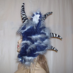 May include: A blue and white faux fur headpiece with black and white striped horns. The headpiece is made of soft, fluffy fur and has a whimsical, fantasy-inspired design.