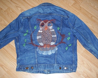 Könnte beinhalten: Eine blaue Jeansjacke mit einem gemalten Eulenmotiv auf der Rückseite. Die Eule ist braun, grau und weiß mit einem großen, blauen Auge. Die Eule sitzt auf einem Ast mit grünen Blättern.