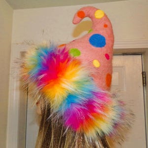 Può includere: Un cappello di feltro rosa con pois colorati e un bordo in pelliccia sintetica arcobaleno. Il cappello ha una corona a punta e un bordo riccio.