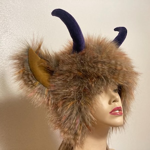 May include: A brown faux fur hat with two curved, dark blue horns and tan-colored ear accents. The hat has a fluffy texture with hints of orange and pink. The hat is on a mannequin head.