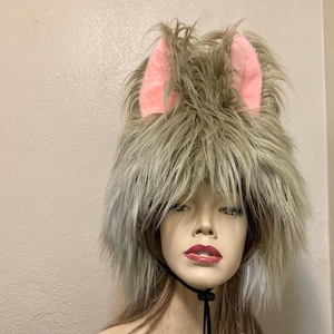 May include: A furry hat with light brown and gray faux fur, featuring two pink, upright ears. The hat has a black chin strap. The hat is on a mannequin head with red lipstick.