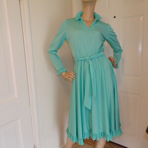 May include: A vintage turquoise blue dress with long sleeves, a ruffled collar, and a matching tie belt. The dress has a flared skirt with ruffles at the hem.