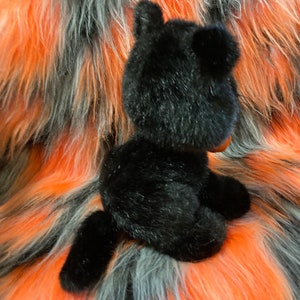 May include: Black plush toy cat sitting on a multi-coloured faux fur background. The cat is facing away from the camera with its tail curled around its body.