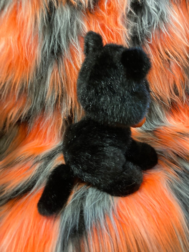 May include: Black plush toy cat sitting on a multi-coloured faux fur background. The cat is facing away from the camera with its tail curled around its body.
