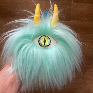 May include: A teal monster plush toy with a large, detailed eye and two yellow horns. The toy has long, shaggy fur and a whimsical design, suitable for imaginative play or as a unique decorative item.