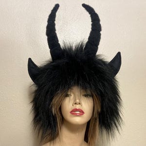 May include: A black faux fur hat featuring two tall, textured, curved horns extending upward from the top and two smaller, pointed, flat-surfaced ears protruding from the sides. The hat is made of long, shaggy black synthetic fur material.