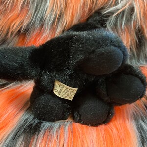 May include: Black plush toy dog with a brown tag that says "Applauds" on it. The dog is lying on a fluffy orange and grey surface.