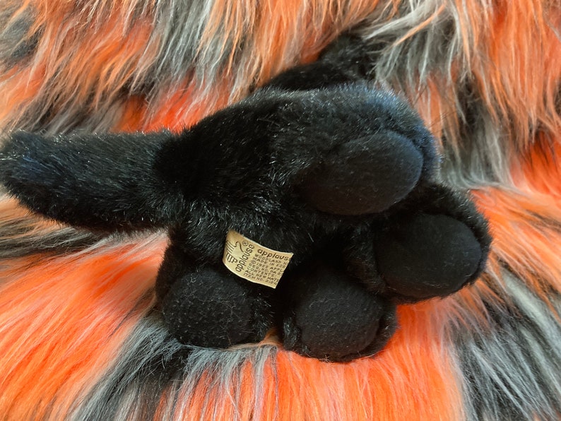 May include: Black plush toy dog with a brown tag that says "Applauds" on it. The dog is lying on a fluffy orange and grey surface.