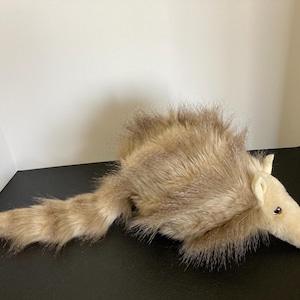 May include: A plush, stuffed aardvark toy. The toy has a long, tubular snout, small, dark eyes, and a long, striped tail. The aardvark's fur is a mix of tan and brown, with a shaggy texture. The toy is lying on a black surface.