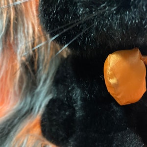 May include: Close-up of a black plush toy with an orange satin bow. The toy has long, fluffy fur.
