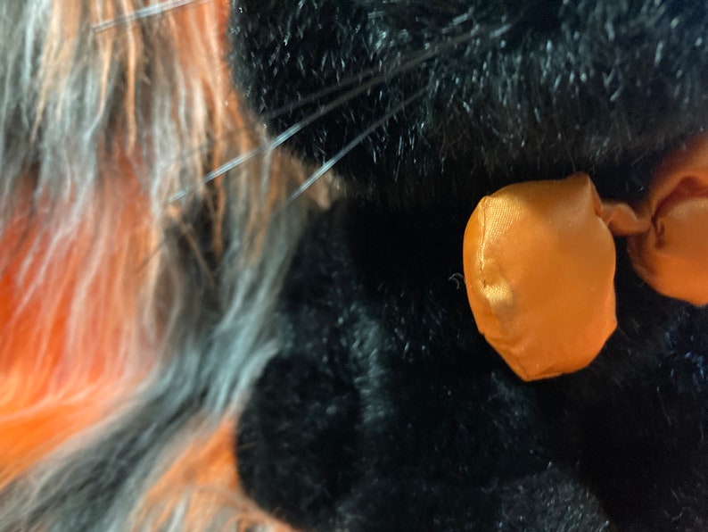 May include: Close-up of a black plush toy with an orange satin bow. The toy has long, fluffy fur.