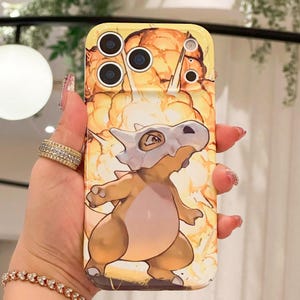 May include: Yellow phone case featuring a cartoon Cubone character with a bone helmet, set against a fiery explosion background. The case has multiple camera cutouts and a glossy finish.