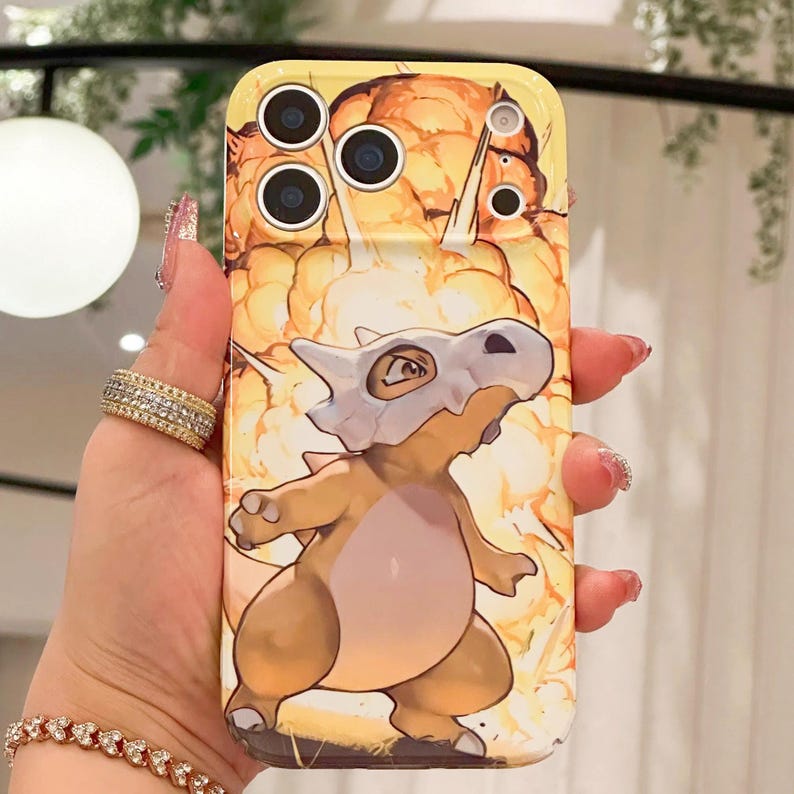 May include: Yellow phone case featuring a cartoon Cubone character with a bone helmet, set against a fiery explosion background. The case has multiple camera cutouts and a glossy finish.