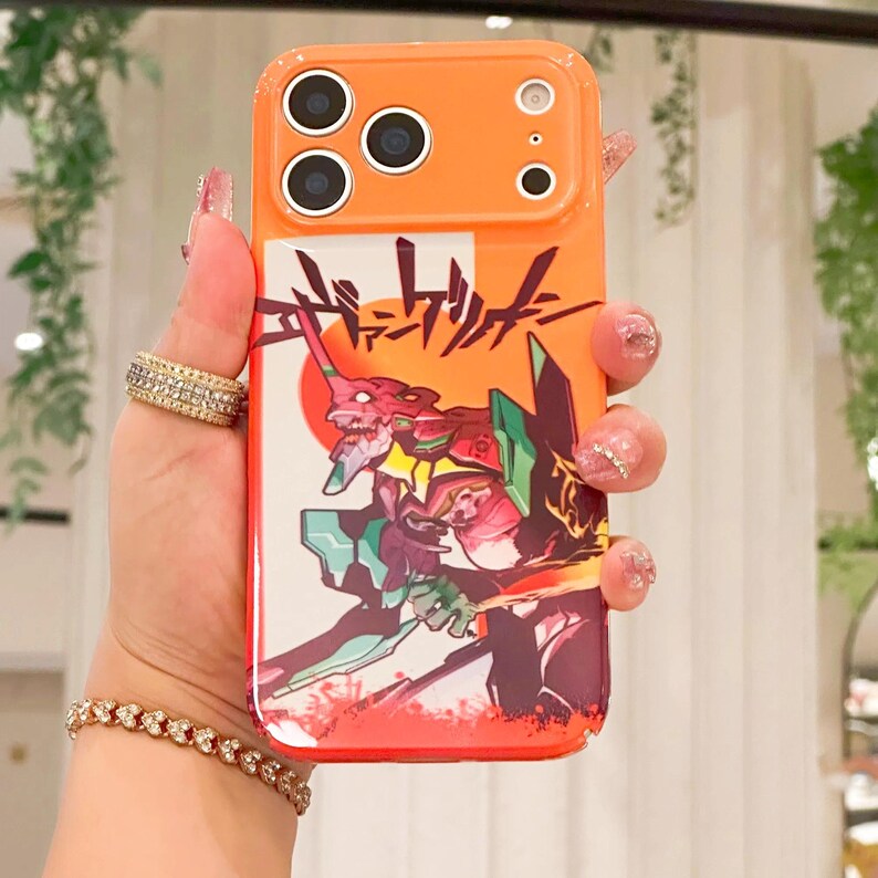 May include: Orange phone case featuring a colorful illustration of an anime character with Japanese text. The case has a camera cutout and is made of a durable material. The design includes red, green, and white elements.