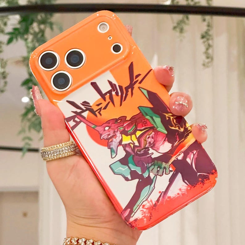 May include: Orange phone case with a graphic of a red and green robot-like figure and Japanese text. The case has a camera cutout and is held in a hand with long, manicured nails and rings.