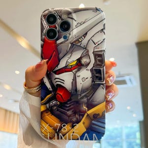 Pu&ograve; includere: Una custodia per telefono bianca, rossa e gialla con un'illustrazione dettagliata di una testa di robot. Il testo "X-78-2 GUNDAM" &egrave; stampato sul fondo della custodia.