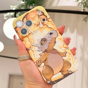 May include: A yellow phone case featuring a cartoon Cubone character with a bone helmet, set against a fiery orange and yellow background. The case has cutouts for the phone's camera and flash.