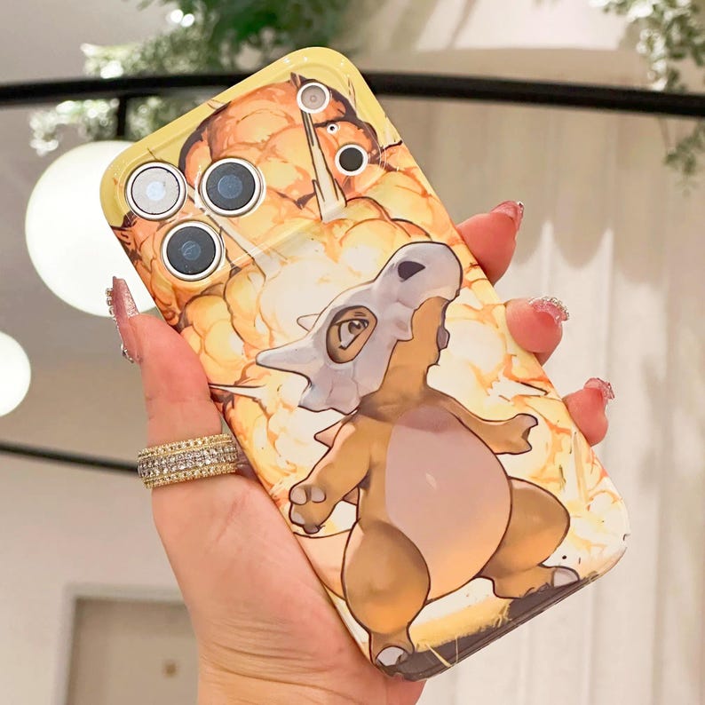 May include: A yellow phone case featuring a cartoon Cubone character with a bone helmet, set against a fiery orange and yellow background. The case has cutouts for the phone's camera and flash.