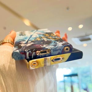Pu&ograve; includere: Due custodie per telefono con un design anime colorato. La custodia superiore ha un design blu, bianco e nero con un bordo dorato. La custodia inferiore ha un design giallo, nero e bianco con un bordo dorato.