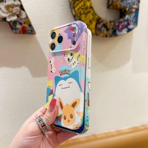 May include: A colorful phone case featuring various Pokemon characters, including Snorlax and Eevee, against a pink and blue background. The case has a glossy finish and includes the word "Pokemon".
