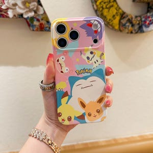 May include: A colorful phone case featuring various Pok&eacute;mon characters. The case has a pink, blue, and yellow background with images of Pikachu, Eevee, Snorlax, and other characters. The word "Pok&eacute;mon" is printed on the case.