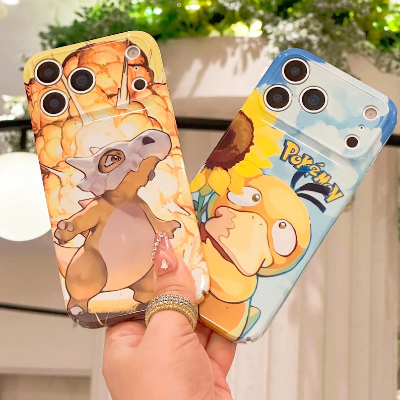 May include: Two phone cases featuring cartoon characters. One case is yellow with a Cubone design, and the other is blue with a Psyduck and sunflower design. The word "Pokemon" is printed on the blue case.