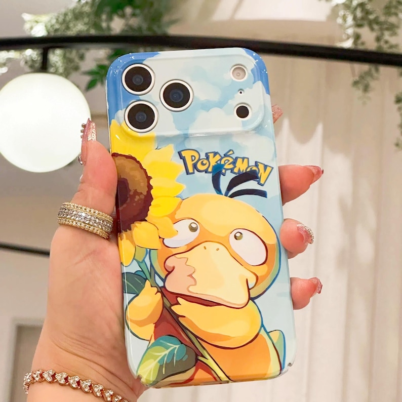 May include: A phone case featuring a cartoon Psyduck character holding a sunflower, set against a blue sky background. The case has the word "Pok&eacute;mon" printed on it. The case is designed to fit a smartphone.