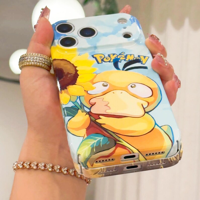 May include: A phone case featuring a cartoon Psyduck character holding a sunflower, set against a blue sky background. The case has the Pokemon logo and a yellow, orange, and blue color scheme.