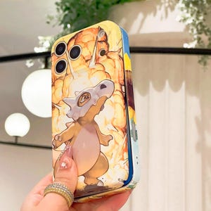 May include: A yellow and blue phone case featuring a cartoon illustration of a Cubone character with a bone helmet, set against a fiery explosion background. The case has a glossy finish and a protective design.