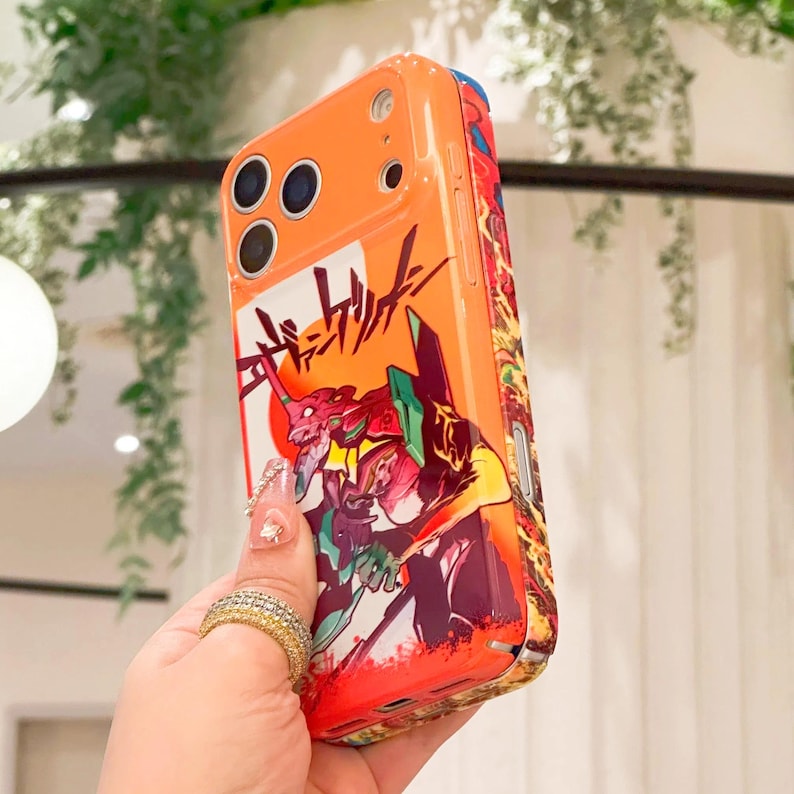 May include: Orange phone case featuring a colorful anime-style illustration with Japanese text. The design includes a red and orange robot-like figure with a rising sun. The case has a glossy finish and a protective design.
