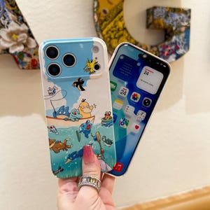 May include: A light blue and white phone case featuring a cartoon illustration of a boat with various characters in the water and sky. The case has a clear camera lens area and is next to a phone with a pink case and a digital display.