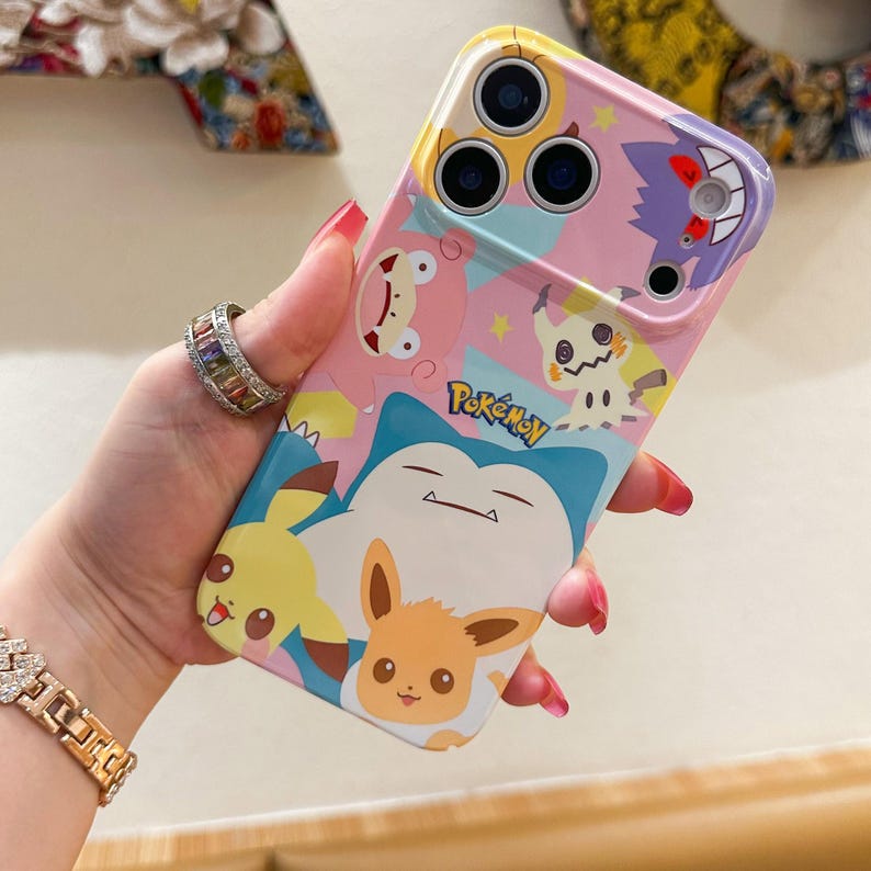 May include: A colorful phone case featuring various Pok&eacute;mon characters, including Pikachu, Eevee, and Snorlax. The case has a pink, blue, and yellow background with the "Pok&eacute;mon" logo. The case is designed to fit a smartphone.