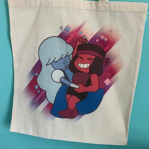 Puede incluir: Una bolsa de tela blanca con una ilustración colorida de dos personajes de dibujos animados. Los personajes son una figura azul y blanca con una diadema roja y una figura roja con una diadema azul. El fondo es un degradado de rosa, morado y azul con formas de diamantes blancos.