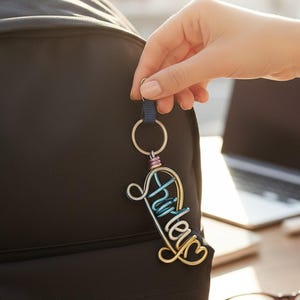 May include: A multi-coloured wire name keyring with the name "Hurley" in a cursive font. The keyring is held by a hand in front of a black backpack. The keyring has a gold, silver, blue, and pink colour scheme.