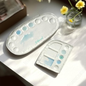 May include: Two white ceramic paint palettes with a cloud design and the names "Dharval" and "Fabiani" in a light green script. The larger palette is oval with circular wells. The smaller palette is rectangular with square and circular wells.