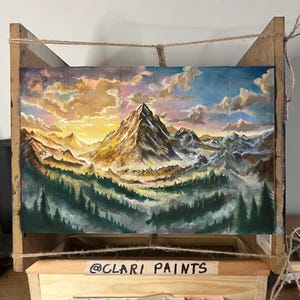 Può includere: Un dipinto che rappresenta una catena montuosa con una cima innevata al tramonto. Il dipinto è realizzato su pannelli di legno e ha un aspetto rustico e vintage. Il nome utente Instagram dell'artista, @CLARI PAINTS, è visibile nella parte inferiore del dipinto.