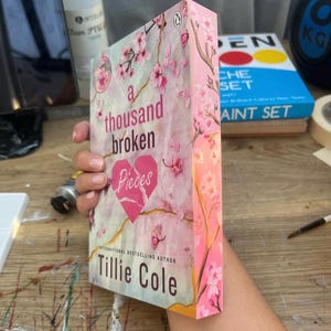 Può includere: Un libro tascabile intitolato "A Thousand Broken Pieces" di Tillie Cole. La copertina presenta uno sfondo in marmo rosa e bianco con un design a forma di cuore spezzato e fiori di ciliegio rosa.