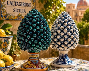 Puede incluir: Dos esculturas de cerámica con forma de piña, una turquesa y otra blanca con detalles azules, sobre una mesa. La piña turquesa tiene una base colorida, mientras que la blanca tiene una base azul y blanca. En el fondo, un letrero que dice "CALTAGIRONE SICILIA".