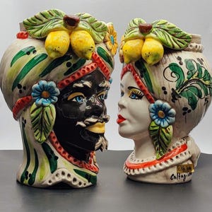 Könnte beinhalten: Zwei Keramikvasen in Kopfform, eine mit schwarzem Gesicht und die andere mit weißem Gesicht. Beide haben farbenfrohe Blumendesigns und Zitronen auf dem Kopf. Die Vase mit dem schwarzen Gesicht hat einen Bart und einen Schnurrbart. Die Vase mit dem weißen Gesicht hat einen rot-weiß gestreiften Turban. Die Vasen sind mit "Caltagirone" signiert.