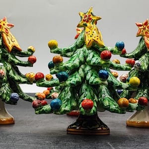 Könnte beinhalten: Eine Sammlung von fünf Keramik-Weihnachtsbaumfiguren. Jeder Baum ist grün mit bunten Ornamenten und einem gelben Stern als Spitze. Die Bäume haben ein detailliertes, handgemaltes Aussehen und stehen auf einer dunklen Oberfläche.