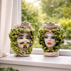 Puede incluir: Dos jarrones de cerámica con forma de cabeza, uno masculino y otro femenino, con rasgos faciales detallados y cabello verde en forma de serpiente. El jarrón masculino tiene bigote y perilla, mientras que la femenina tiene labios rojos. Ambos tienen ojos azules y están decorados con detalles crema y verde.