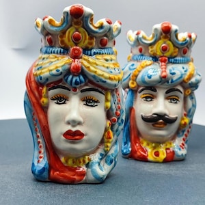 Könnte beinhalten: Zwei Keramikfiguren mit stilisierten Gesichtern und Kronen, eine mit einem rot-blauen Turban und die andere mit einem blau-gelben Turban. Beide haben detaillierte Gesichtszüge und farbenfrohe Akzente.