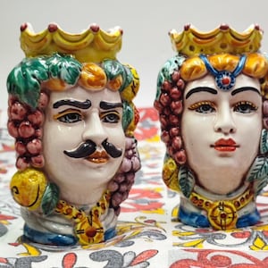 May include: Two ceramic head vases, one male and one female, with detailed facial features and ornate crowns. The male vase has a mustache and is adorned with grapes and lemons. The female vase has a necklace and is decorated with grapes.